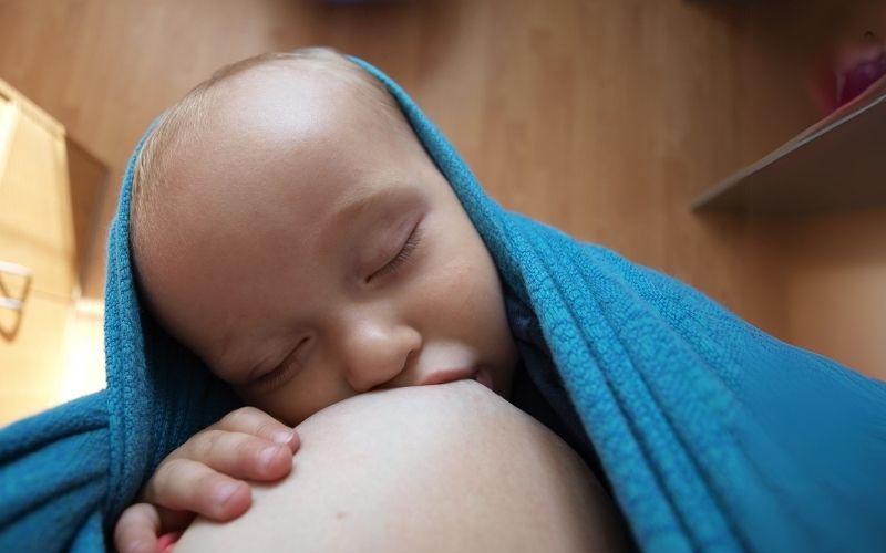 Bábätko zabalené v modrej šatke spokojne pije materské mlieko počas dojčenia – pokojná a láskyplná chvíľa spojenia s mamou