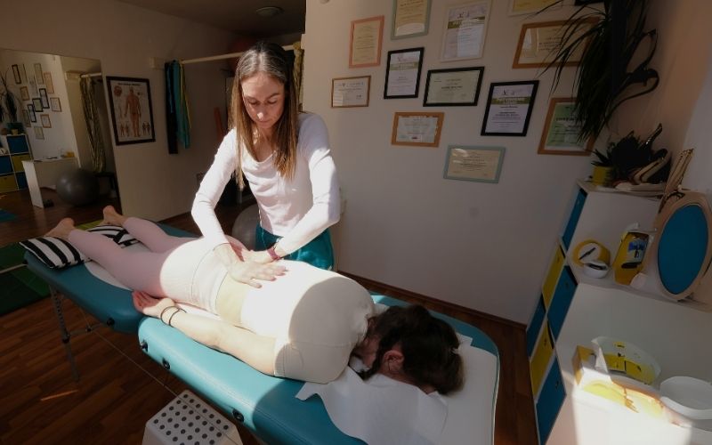 Fyzioterapeutka vykonáva terapeutickú masáž na klientke ležiacej na masážnom stole v svetlej a útulnej miestnosti plnej certifikátov.