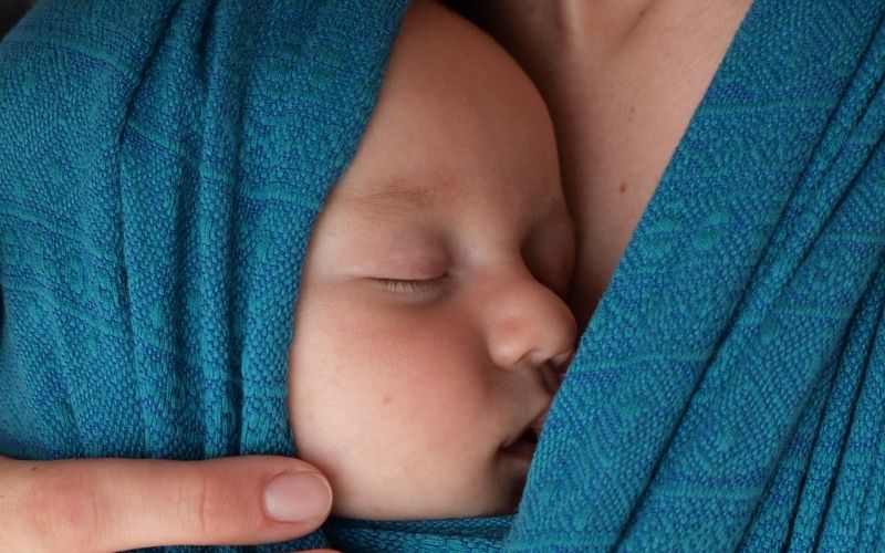Spiace bábätko zabalené v modrej šatke spočíva na hrudi mamy – moment pokoja, bezpečia a blízkosti.