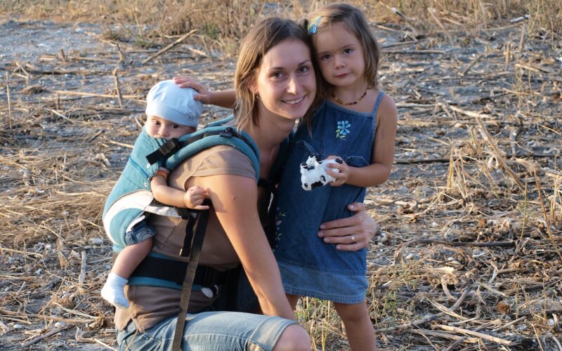mamka s dvomi deťmi, jedno v nosiči na chrbte a druhé vedľa nej
