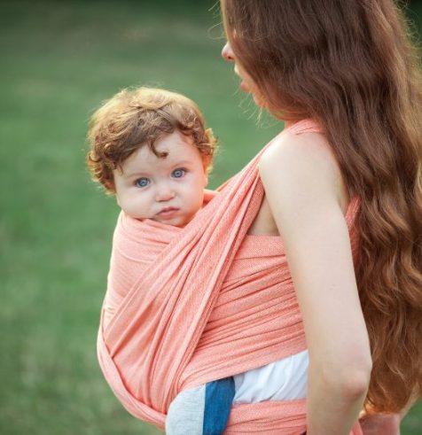 Mama s dlhými vlasmi nosí dieťa v korálovo-oranžovej šatke na nosenie v prírode. Dieťa je pokojné, pritúlené k mame a s dôverou sa pozerá na svet.